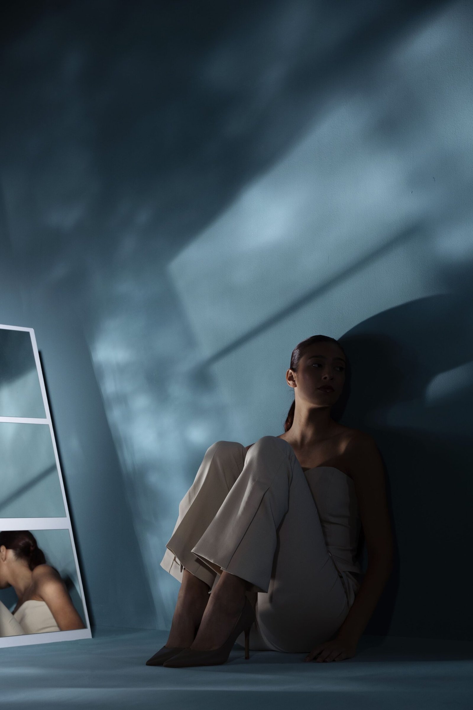 A woman sits on the floor against a dimly lit blue wall, her knees drawn close to her chest, gazing downward in deep contemplation. Soft shadows and reflections in a nearby mirror enhance the somber, introspective mood visually expressing the weight of anxiety from past trauma and emotional isolation.