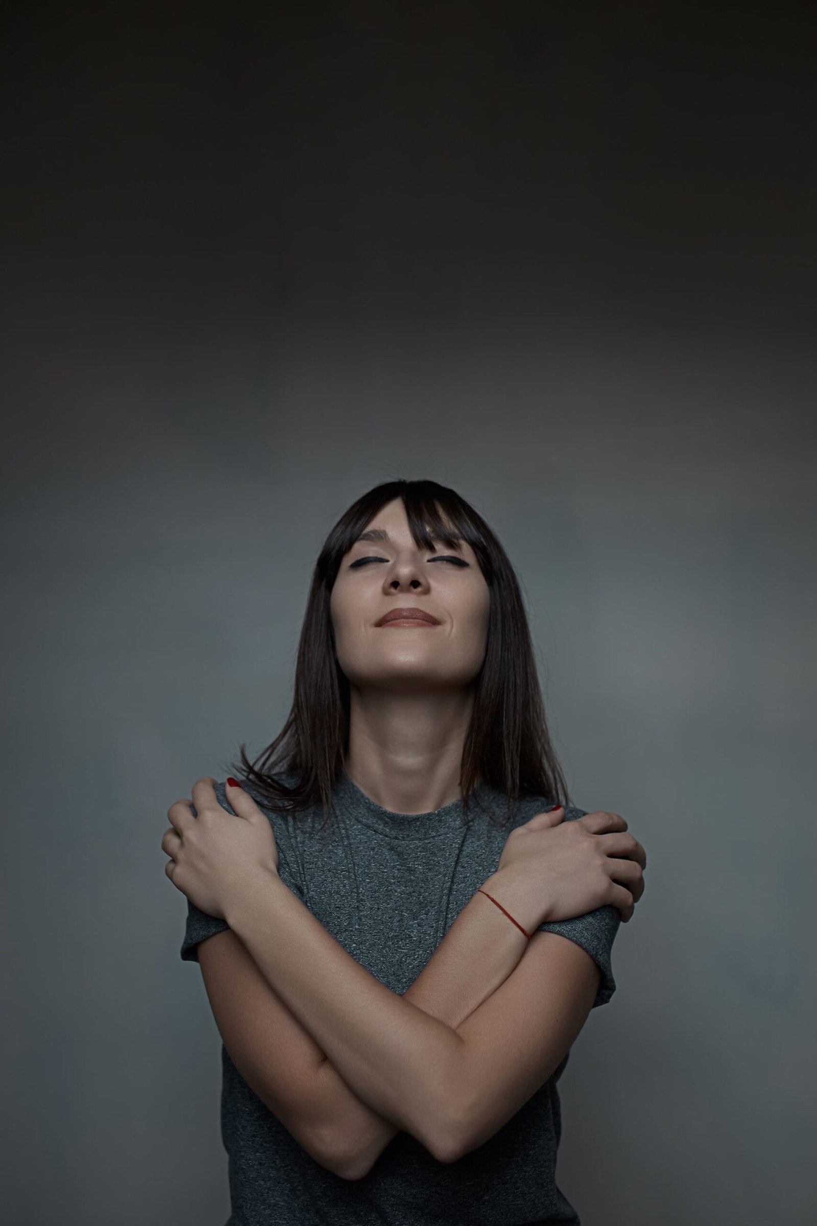 A woman stands with her eyes closed, gently hugging herself and smiling softly, radiating calm and self-acceptance. The serene expression and neutral background evoke a sense of healing, inner peace, and emotional recovery from anxiety from past trauma.