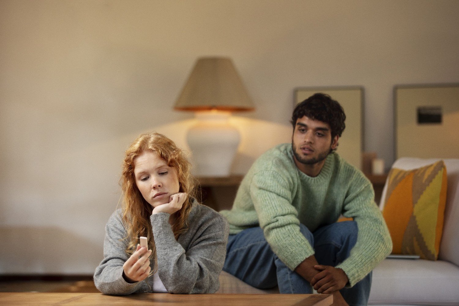A couple sitting apart in a living room, the woman looking down sadly while the man watches her with concern, representing guilt in relationships and emotional distance.