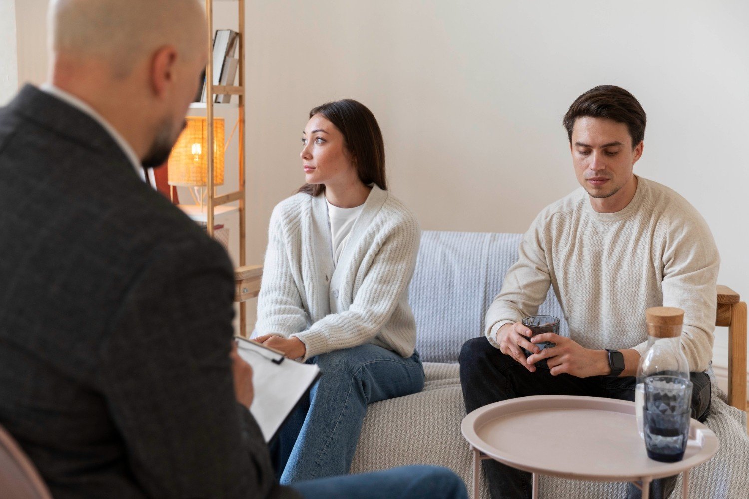 A couple sits with a therapist during a counseling session, both appearing distant and reflective, representing couples therapy after infidelity and efforts to rebuild trust.