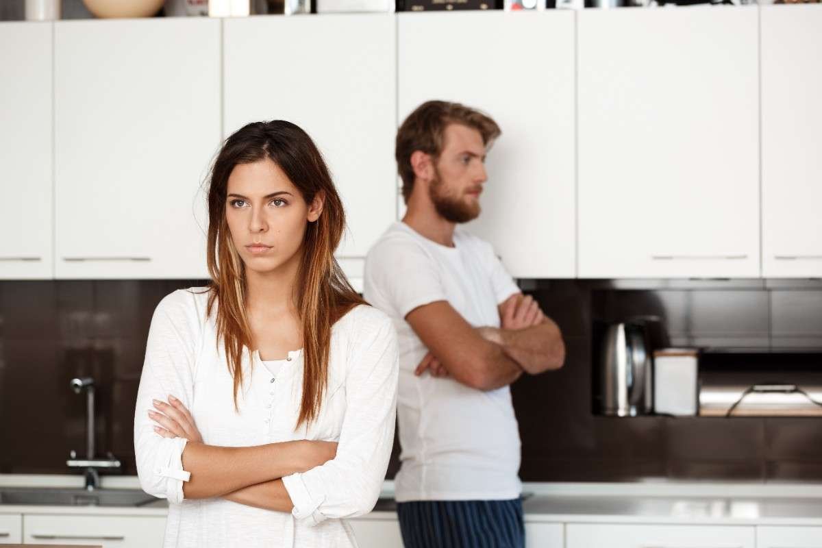 A couple standing apart in a modern kitchen with crossed arms and tense expressions, reflecting the challenge of how to communicate without arguing during relationship conflicts.