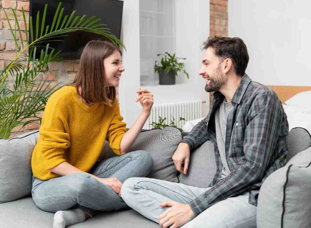 A couple sitting comfortably on a sofa, smiling and talking openly in a relaxed home setting, showing healthy relationship communication and emotional connection.