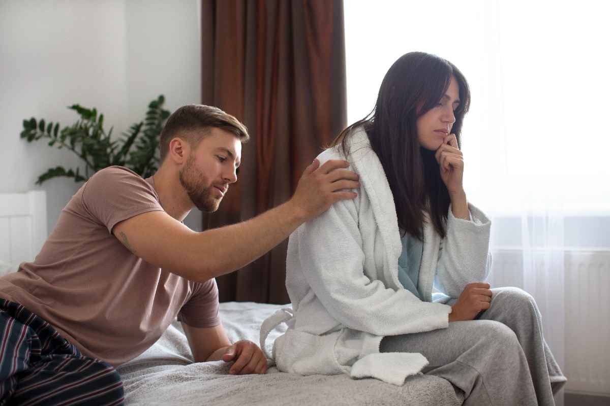 A man gently trying to comfort his partner sitting quietly on the bed, showing emotional distance and early signs you need couple counselling in a relationship.