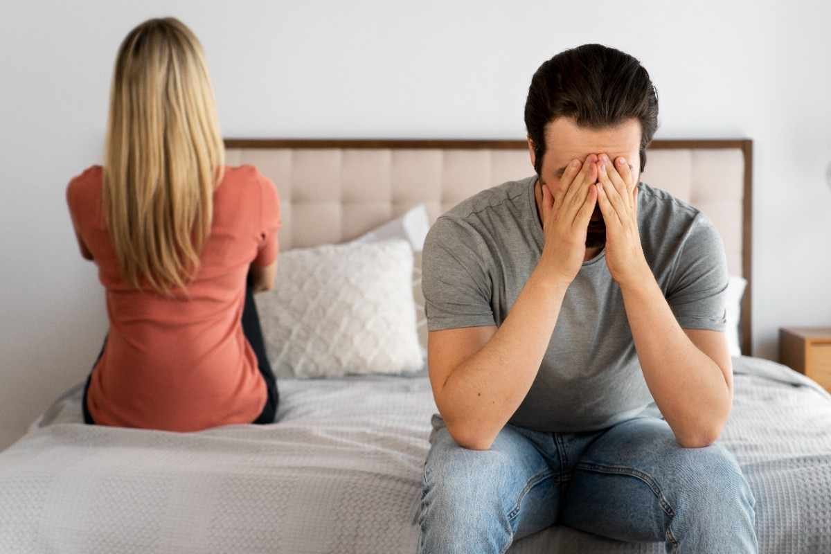 A man sitting on the bed with his head in his hands while his partner turns away, showing emotional strain and signs you need couple counselling in a struggling relationship.